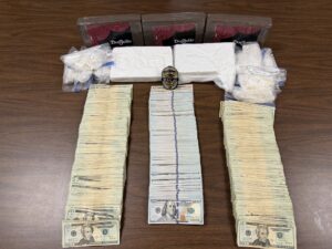 A large display of seized items arranged on a wooden table. At the top are several rectangular, shrink‑wrapped packages labeled “Don Julio.” In front of them are two large white rectangular blocks with embossed textures, positioned among multiple smaller sealed plastic bags containing white, powdery or rock‑like substances. A law‑enforcement badge is placed at the center of the arrangement. Three tall stacks of bundled U.S. currency are lined up below the packages, with denominations visible on the top of each stack.