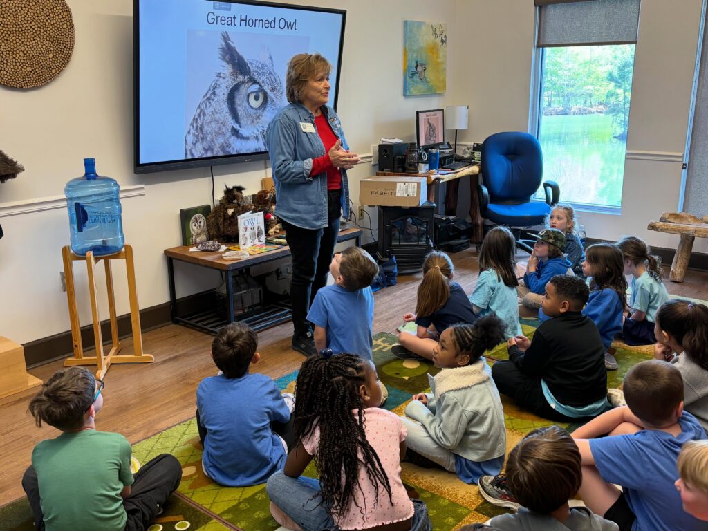 Elementary students sit on the floor in a classroom listening to a Green Team ambassador talk about the importance of owls in nature.