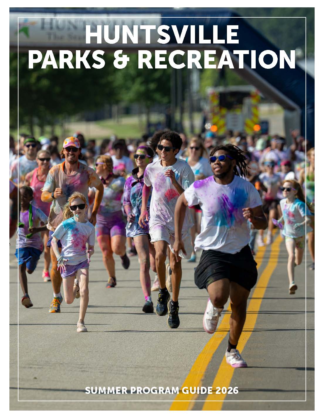 A large group of people of various ages run down a city street during a colorful fun‑run event, their white shirts covered in bright paint powder. A starting archway and trees line the background. Bold text at the top reads “Huntsville Parks & Recreation,” and text at the bottom reads “Summer Program Guide 2026.”