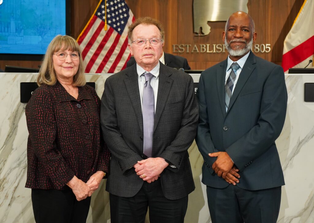 a women on left and two men in suits pose for photo with flags in background