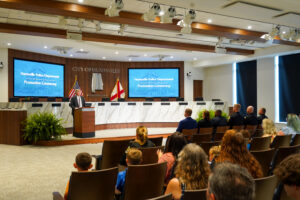 A promotion ceremony taking place inside a formal meeting chamber. A speaker stands at a podium at the front of the room, with large display screens behind showing the text “Huntsville Police Department Promotion Ceremony.” The backdrop features the “City of Huntsville” emblem along with two flags positioned behind the podium. Rows of attendees sit facing the front, including uniformed personnel and family members. The room features wood accents, marble surfaces, and ceiling-mounted lighting.