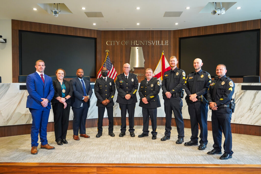 A group of individuals in professional attire and police uniforms stand in a row inside a formal meeting chamber. The backdrop features a large wooden panel with the text “City of Huntsville” and two flags positioned behind the group, including the United States flag and another government flag. The room includes marble accents, a raised dais, and large display screens on both sides. The group is posed facing the camera for a formal photo.