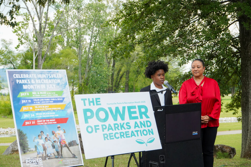 A young man speaks into a microphone at a podium and woman standing near observes. Next to the podium are decorative signs and the background is a park.