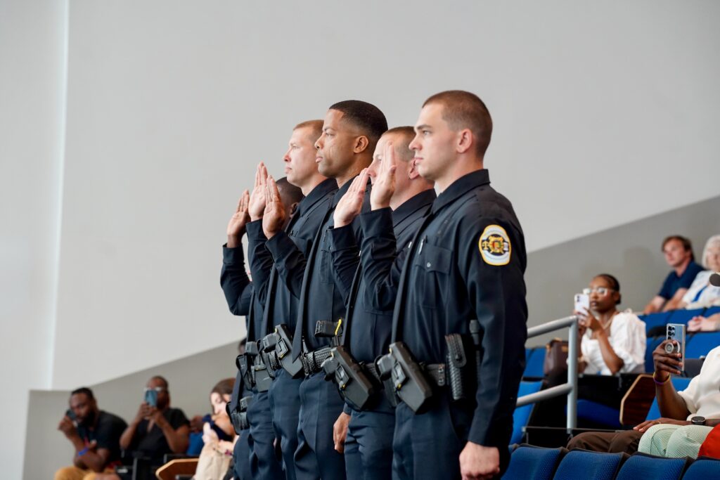 Four uniformed officers stand in a row with their right hands raised during a formal ceremony. They wear dark uniforms with visible badges and equipment. In the background, people are seated in an auditorium, some taking photos or observing the event.