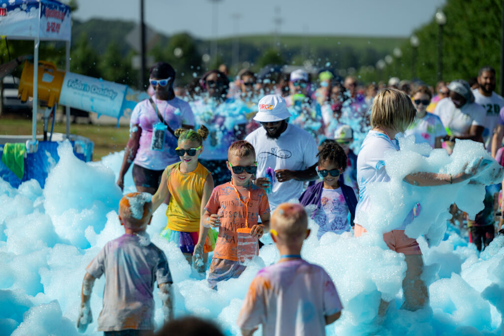 Children and adults wearing colorful shirts and sunglasses walk through a large area filled with blue-tinted foam during the Color Jam Family Run & Walk at John Hunt Park. Participants smile and play in the foam while a crowd gathers in the background on a sunny day.