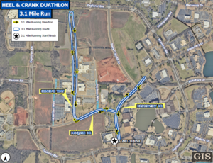 An aerial map shows the 3.1‑mile running route for the Heel & Crank Duathlon. The course is marked with a blue line and yellow arrows indicating the running direction. The path follows several roads, including Genome Way, Moquin Drive, Discovery Drive, and sections near Mark C. Smith Drive and Hudson Way. A star icon marks the start and finish area near the lower center of the map. A legend in the upper left corner explains the symbols for route, direction, and start/finish. The background displays buildings, parking lots, open fields, and surrounding roadways, with a GIS City of Huntsville logo in the bottom right corner.