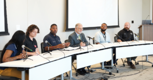 A panel of six individuals is seated at a long white table with microphones in front of each person. The individuals are diverse and participating in a media roundtable discussion.
