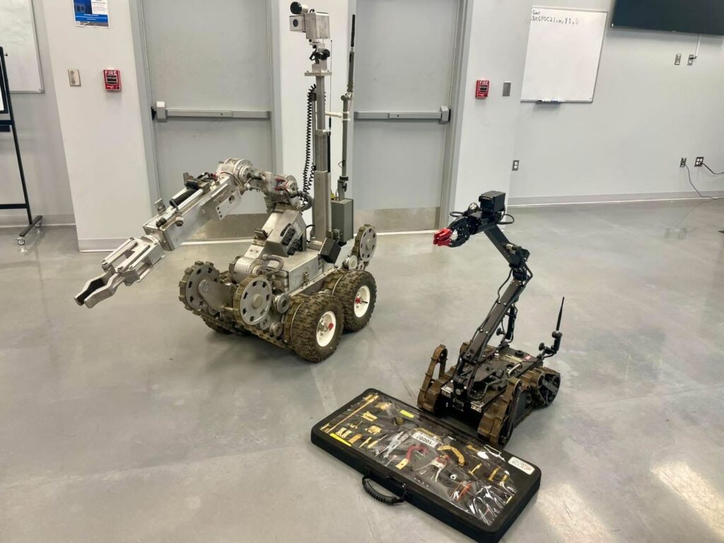 Two robotic vehicles are positioned in a room with a gray floor and white walls. The larger robot on the left has a mechanical arm and is equipped with treads. The smaller robot on the right is also equipped with an arm and treads. A toolkit is displayed on the floor in front of them. In the background, there are closed double doors, a whiteboard, and a black stand.