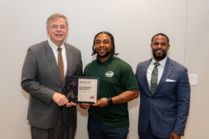 Three people standing together as a Civic Engagement Academy graduate holds a framed certificate of participation; one person stands to the graduate’s left wearing a suit and another to the right also wearing a suit.