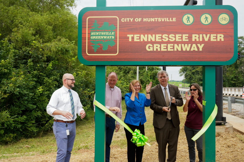 Five individuals stand in front of the Tennessee River Greenway sign in Huntsville during a ribbon‑cutting ceremony. One person cuts a green ribbon while the others hold it. The sign features the Huntsville Greenways logo and icons for walking and bicycling. Trees and part of a roadway are visible behind the sign.