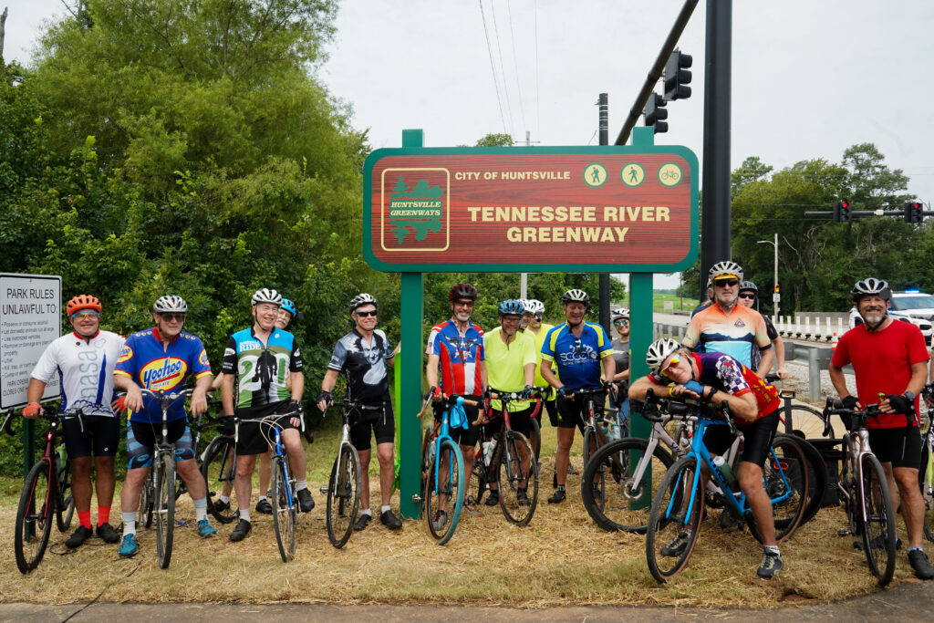 A large group of cyclists pose with their bicycles in front of the Tennessee River Greenway sign in Huntsville. They are standing together on a mulched area next to the paved trail. The greenway sign features walking and cycling icons and the Huntsville Greenways logo. Trees, traffic lights, and a roadway are visible in the background.