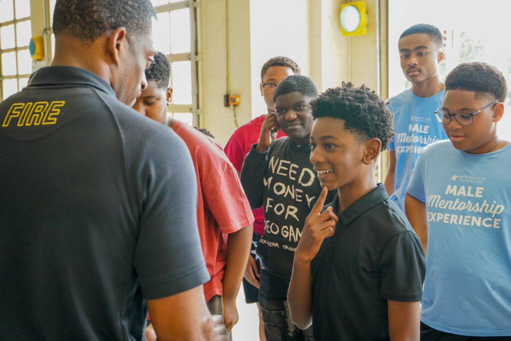 A Huntsville Fire & Rescue firefighter speaks with participants in the Huntsville Parks & Recreation Male Mentorship Experience during a fire station visit. Several boys listen as one participant smiles and asks a question while others look on.