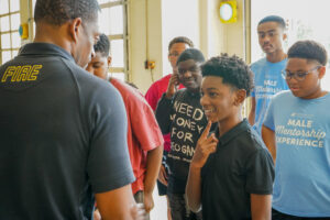 A Huntsville Fire & Rescue firefighter speaks with participants in the Huntsville Parks & Recreation Male Mentorship Experience during a fire station visit. Several boys listen as one participant smiles and asks a question while others look on.