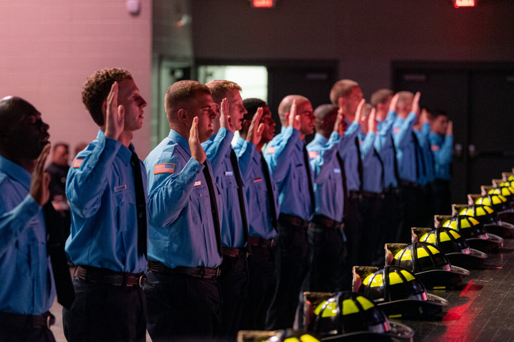 Row of firefighters in blue shirts with right arms raised at graduation