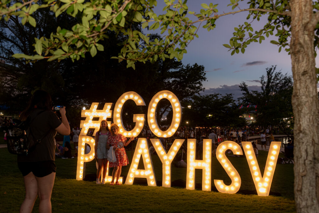 Two people pose for a photo in front of a large illuminated "#GoPlayHSV" sign at Big Spring Park during an evening community event while another person takes their picture.