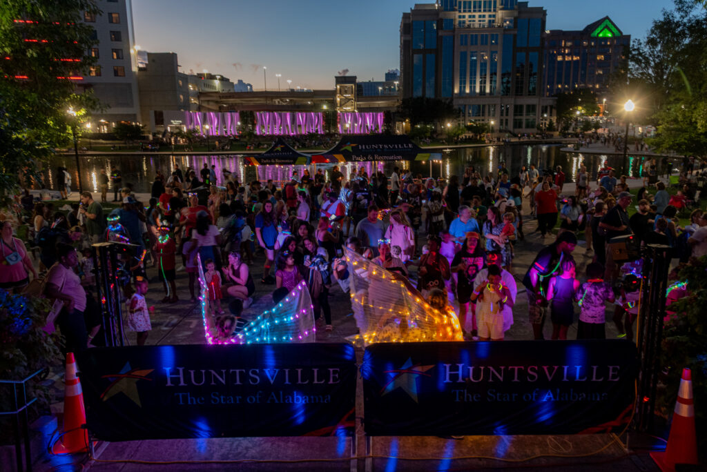 A large crowd gathers at Big Spring Park during a nighttime community event. People wearing glow accessories fill the plaza near the water while illuminated performers and the Huntsville skyline create a colorful backdrop