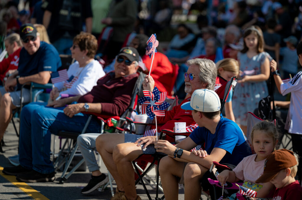 People sitting in chairs watching an event, many holding American flags and patriotic decorations.