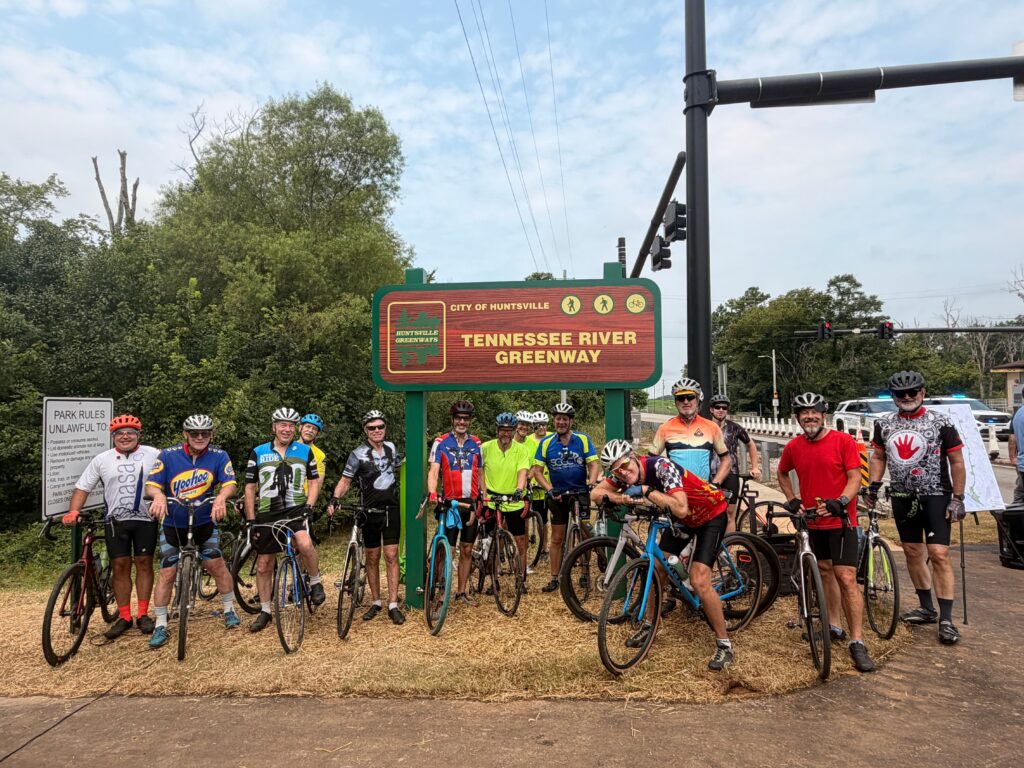 A large group of cyclists pose with their bicycles in front of the Tennessee River Greenway sign in Huntsville. They are standing together on a mulched area next to the paved trail. The greenway sign features walking and cycling icons and the Huntsville Greenways logo. Trees, traffic lights, and a roadway are visible in the background.