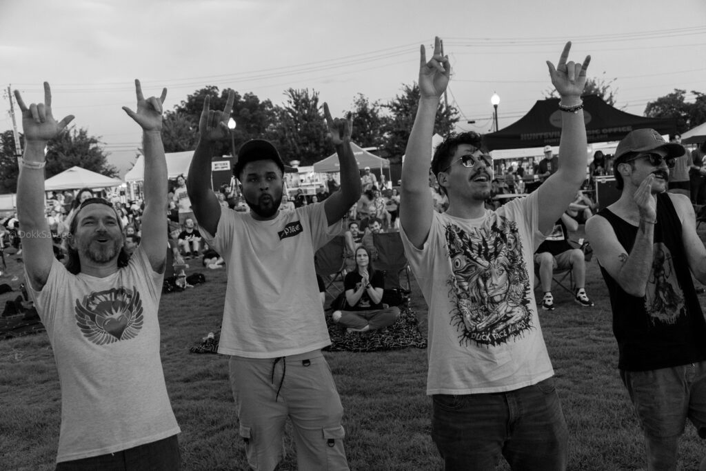 A group of people at an outdoor concert stand on the grass with their arms raised, making rock‑and‑roll hand signs. Tents, seated attendees, and event booths are visible in the background under an evening sky.