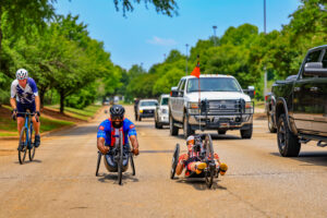 The image shows several athletes participating in a road cycling event, including two athletes using handcycles and another athlete riding a traditional bicycle. Multiple vehicles follow behind them on a tree‑lined street under bright, sunny conditions. Photo credit: Huntsville/Madison County Chamber.