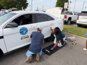 Two people kneel beside a white City of Huntsville Motor Pool vehicle, working near the front wheel with tools and a kneeling mat on the ground. The vehicle’s hood is open, and other parked vehicles and an 'Employee Parking Only' sign are visible in the background.