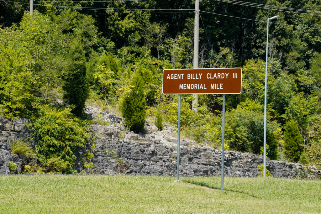 The image shows a brown roadside sign standing on metal posts in front of a rocky embankment and dense green foliage. The sign reads “Agent Billy Clardy III Memorial Mile” in white capital letters. Grass covers the foreground, and utility poles and wires are visible above the trees in the background.