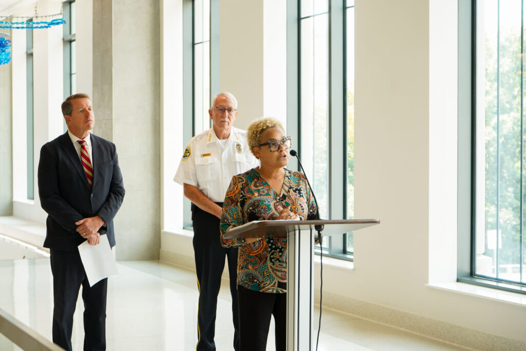 A person stands at a podium speaking into a microphone in a bright hallway lined with tall windows. Two other individuals stand behind the speaker, one holding papers and the other wearing a uniform-style shirt. The setting features polished floors, large window panels, and natural light illuminating the space.