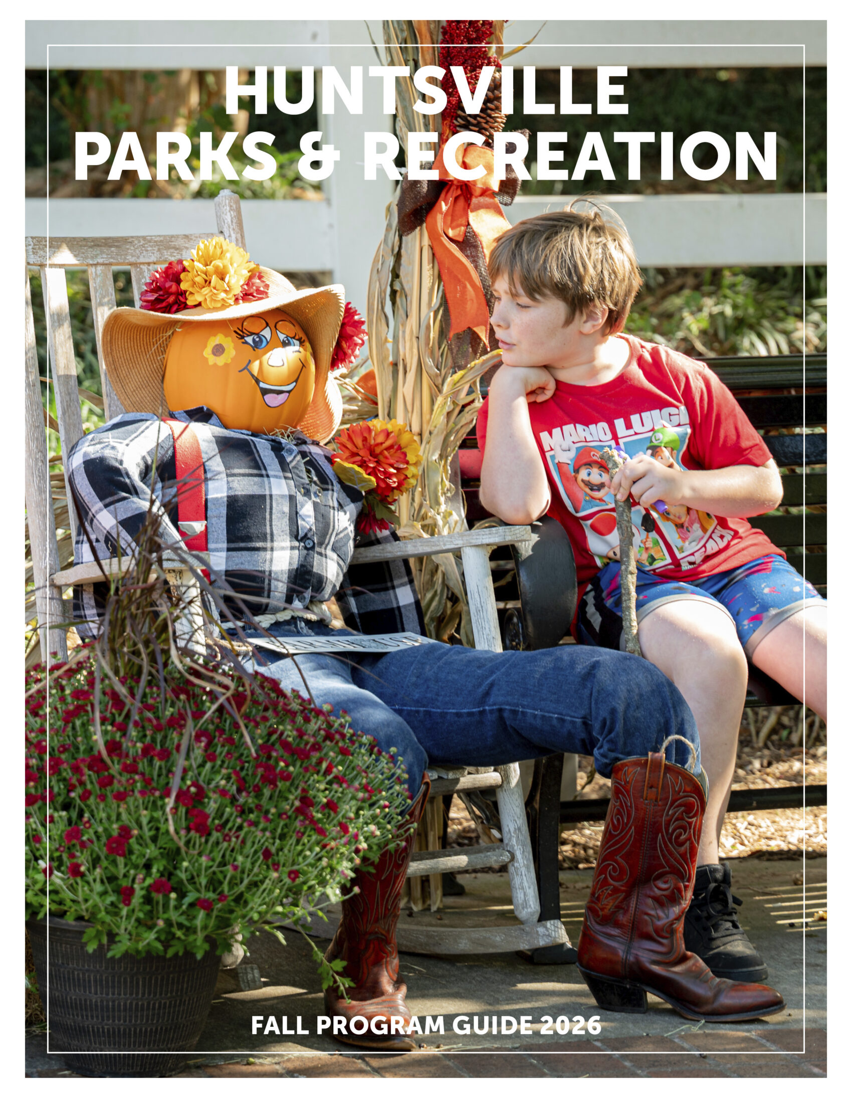 A fall-themed cover for the Huntsville Parks & Recreation Fall Program Guide 2026. The image shows a seated decorative scarecrow with a pumpkin head, wearing a plaid shirt, jeans, cowboy boots, and a straw hat adorned with flowers. A child sits nearby on a wooden bench, leaning toward the scarecrow while holding a stick. Behind them are corn stalks, flowers, and a white fence. Text at the top reads “Huntsville Parks & Recreation,” and text at the bottom reads “Fall Program Guide 2026.”