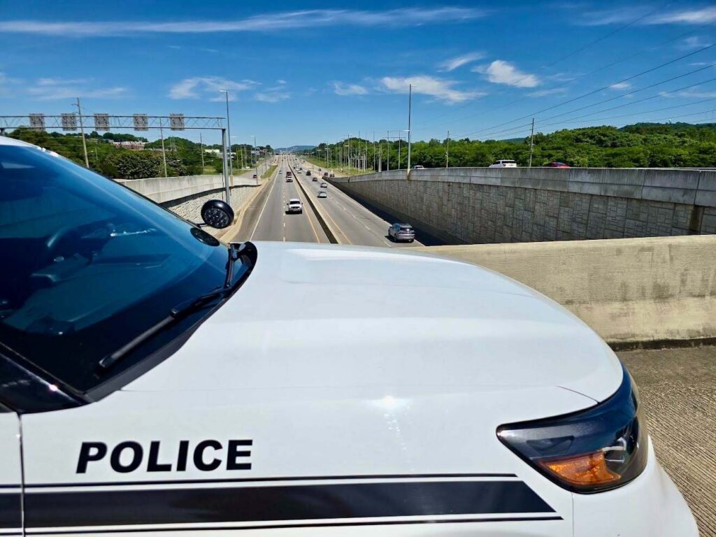 A partial view of a marked police vehicle parked on an overpass, overlooking a multi-lane highway with light traffic traveling in both directions. The highway is lined with concrete retaining walls, utility poles, and green tree-covered hills under a bright blue sky with scattered clouds.