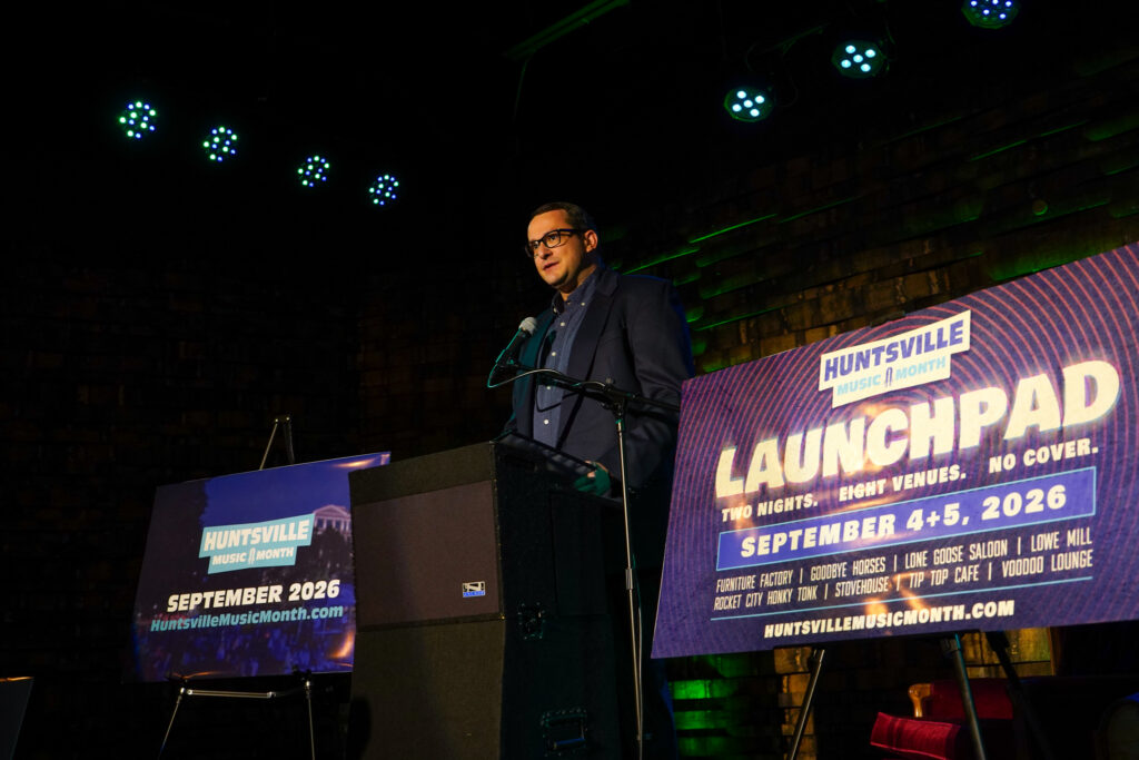 man in dark shirt and jacket speaks at podium with posters on each side