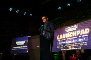 man in dark shirt and jacket speaks at podium with posters on each side