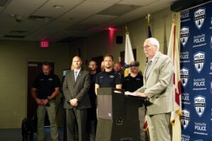 The image shows an indoor press conference setting where an individual in a suit is speaking at a podium with a microphone. Behind the speaker, several uniformed individuals stand in a row, along with a dog on a leash held by another uniformed individual. A blue backdrop on the right displays multiple “Huntsville Police” logos, and various flags are positioned near the podium.