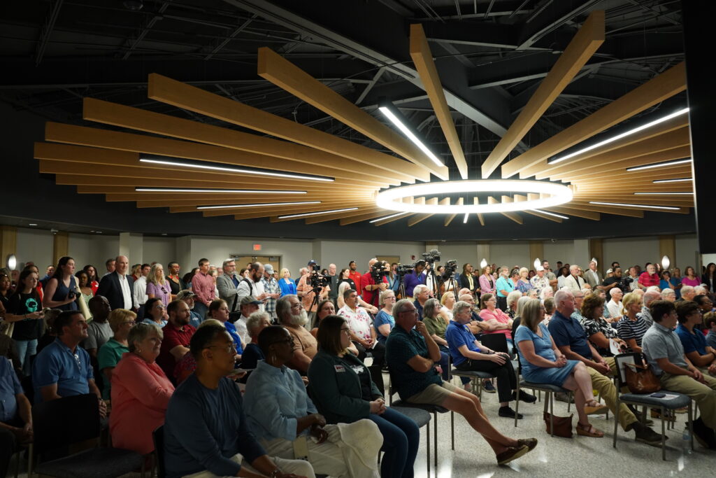 A crowd sitting and standing in an event space with overhead lighting and wood paneling.