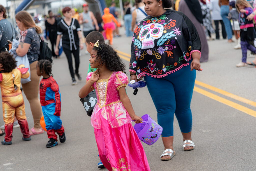 Children and adults in Halloween costumes walk through a community event, including a child dressed as a princess and another dressed as Spider-Man.