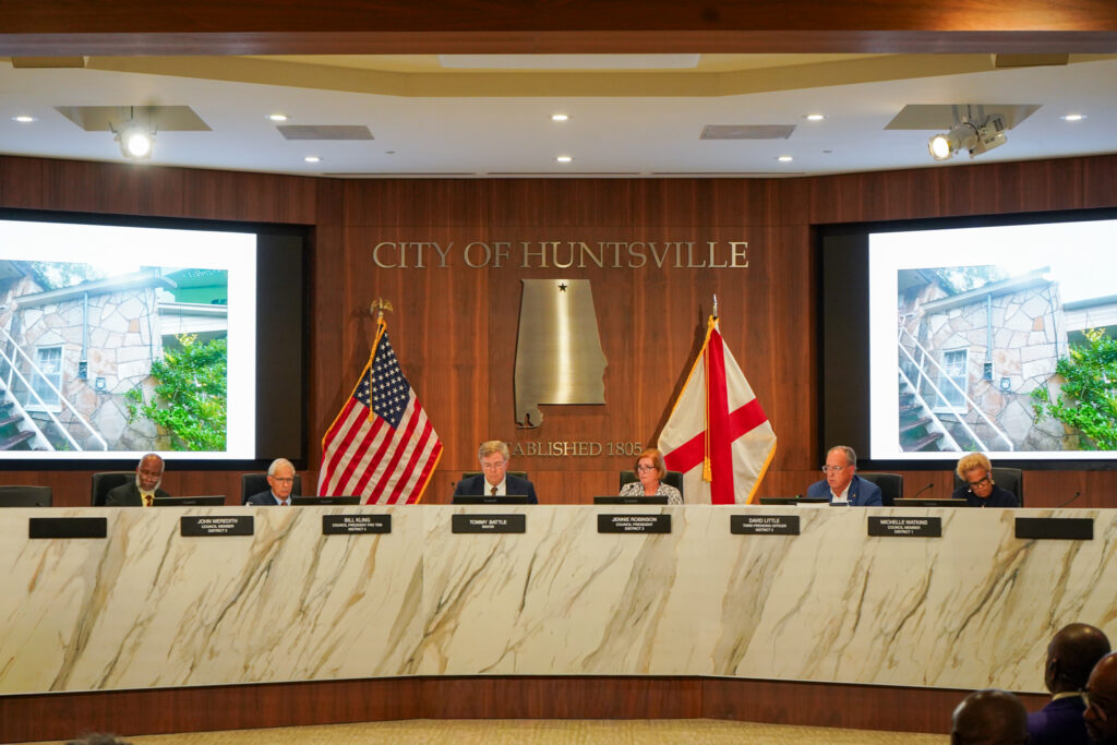 mayor and city council seated at white dais with flags and projection screens in background