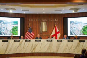 mayor and city council seated at white dais with flags and projection screens in background