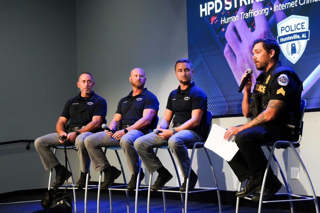 Four individuals sit on tall chairs on a stage, each holding a microphone or notes, while participating in a panel discussion. A large screen behind them displays the title “HPD Strike Unit: Human Trafficking • Internet Crimes Against Children,” and a working dog is lying on the floor in front of the panel.